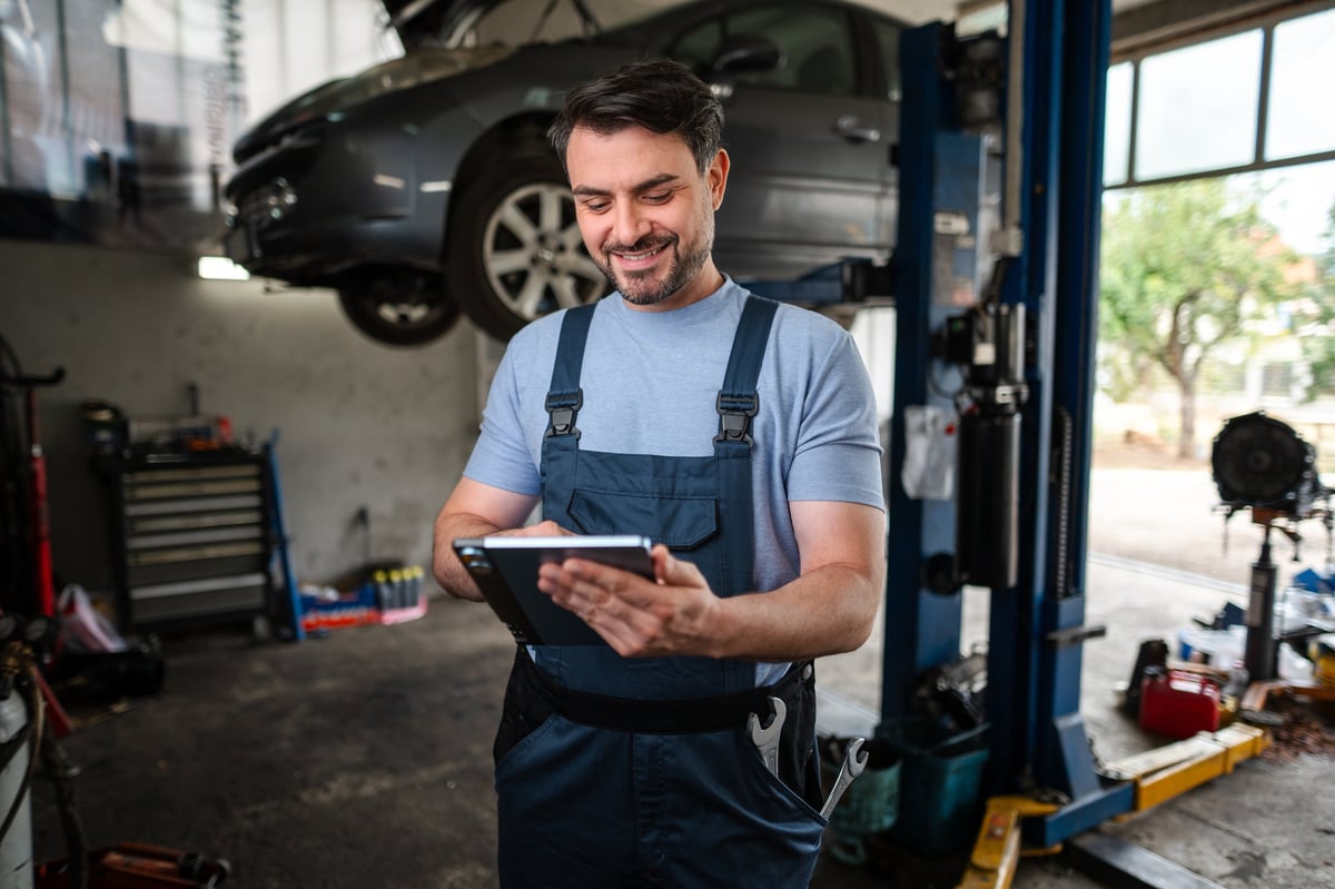 Smiling mechanic using a digital tablet next to a car on a hydraulic lift in a auto repair shop